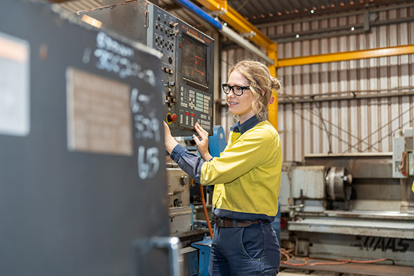 Woman with blonde hair, wearing black rimmed glasses and high visibility clothing stands in front of a machine control board with lots of buttons